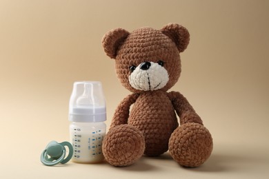 Photo of Feeding bottle with milk, pacifier and toy bear on beige background