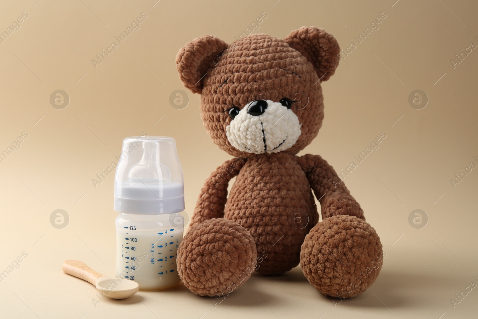 Photo of Feeding bottle with milk, spoon and toy bear on beige background