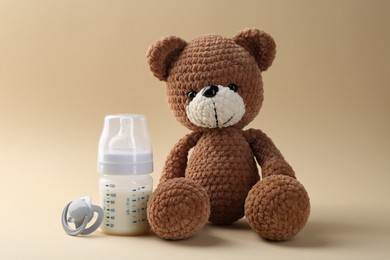 Photo of Feeding bottle with milk, pacifier and toy bear on beige background