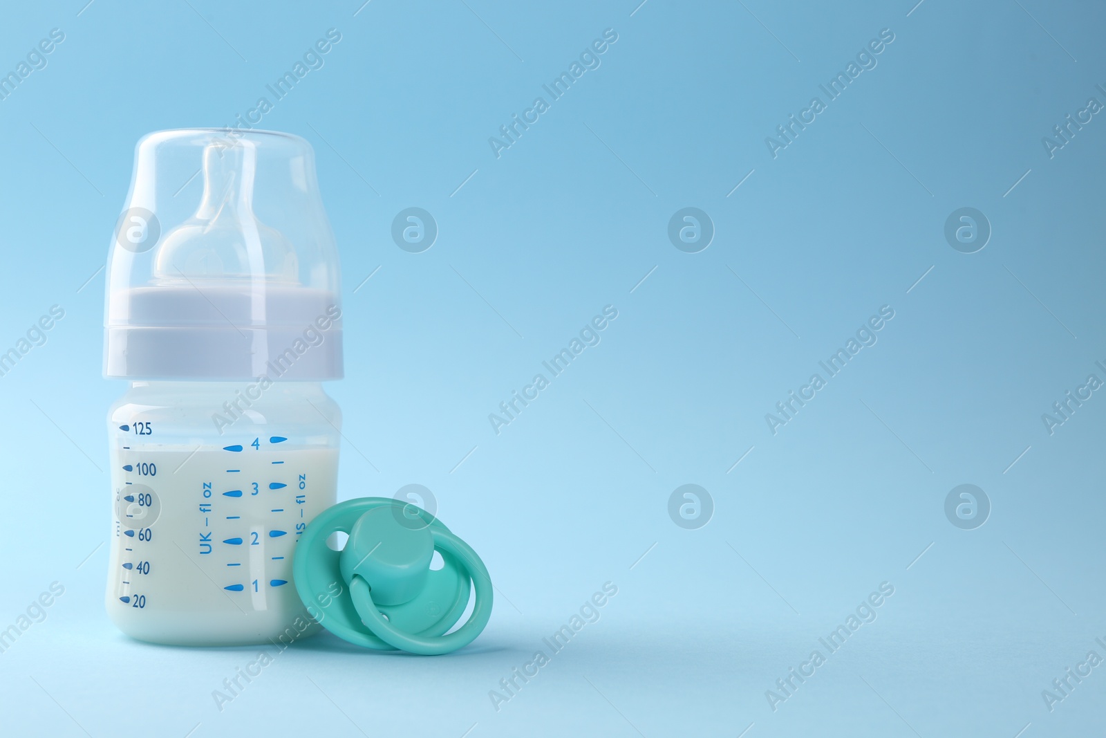 Photo of Feeding bottle with milk and pacifier on light blue background, space for text