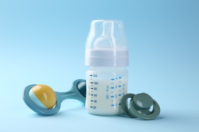 Photo of Feeding bottle with milk, pacifier and baby rattle on light blue background