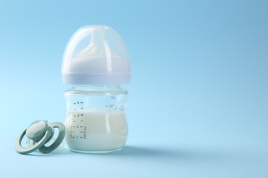 Feeding bottle with milk and pacifier on light blue background, space for text