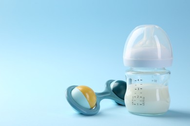 Photo of Feeding bottle with milk and baby rattle on light blue background, space for text