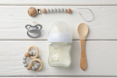 Feeding bottle with milk, spoon, pacifier and teethers on white wooden table, flat lay