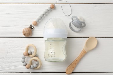 Feeding bottle with milk, spoon, pacifier and teethers on white wooden table, flat lay