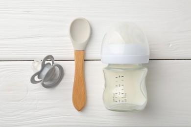 Feeding bottle with milk, spoon and pacifier on white wooden table, flat lay