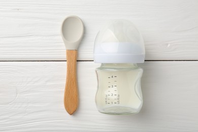 Feeding bottle with milk and spoon on white wooden table, flat lay