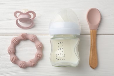 Feeding bottle with milk, spoon, pacifier and teether on white wooden table, flat lay