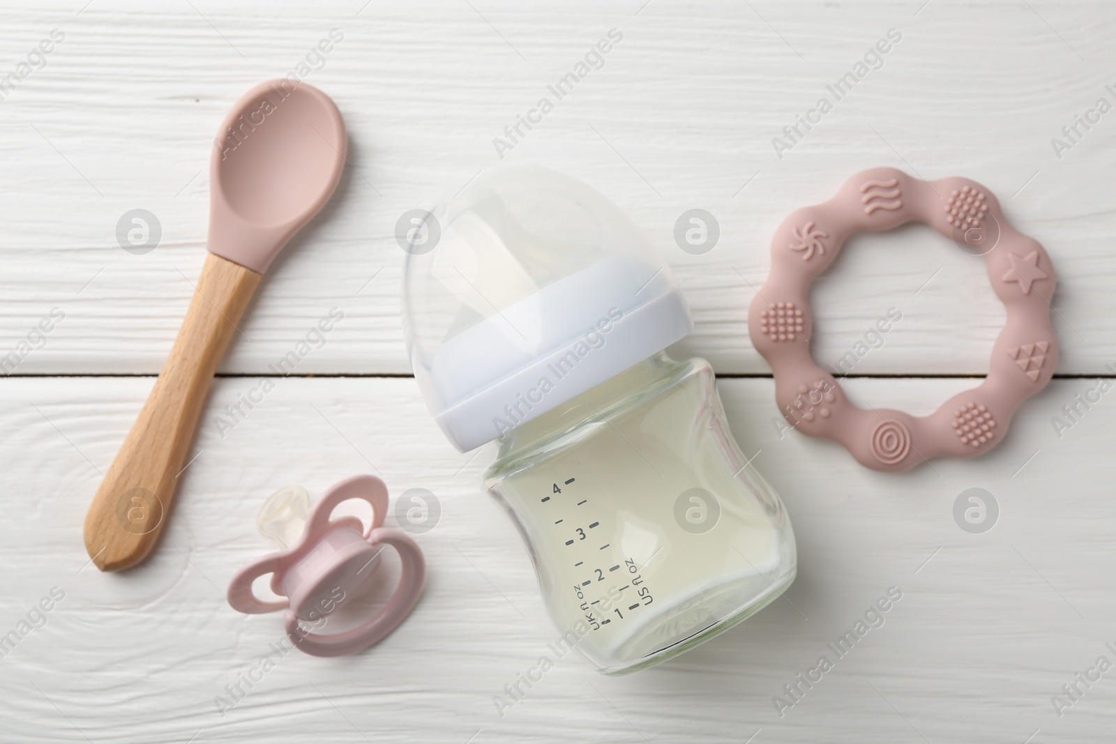 Photo of Feeding bottle with milk, spoon, pacifier and teether on white wooden table, flat lay