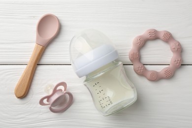 Feeding bottle with milk, spoon, pacifier and teether on white wooden table, flat lay