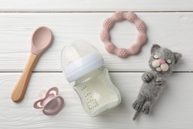 Feeding bottle with milk, needle felted cat, pacifier, teether and spoon on white wooden table, flat lay