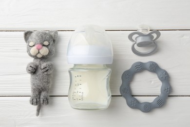 Feeding bottle with milk, needle felted cat, pacifier and teether on white wooden table, flat lay