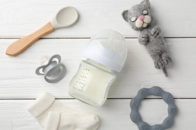 Feeding bottle with milk, needle felted cat, pacifier, spoon, baby socks and teether on white wooden table, flat lay