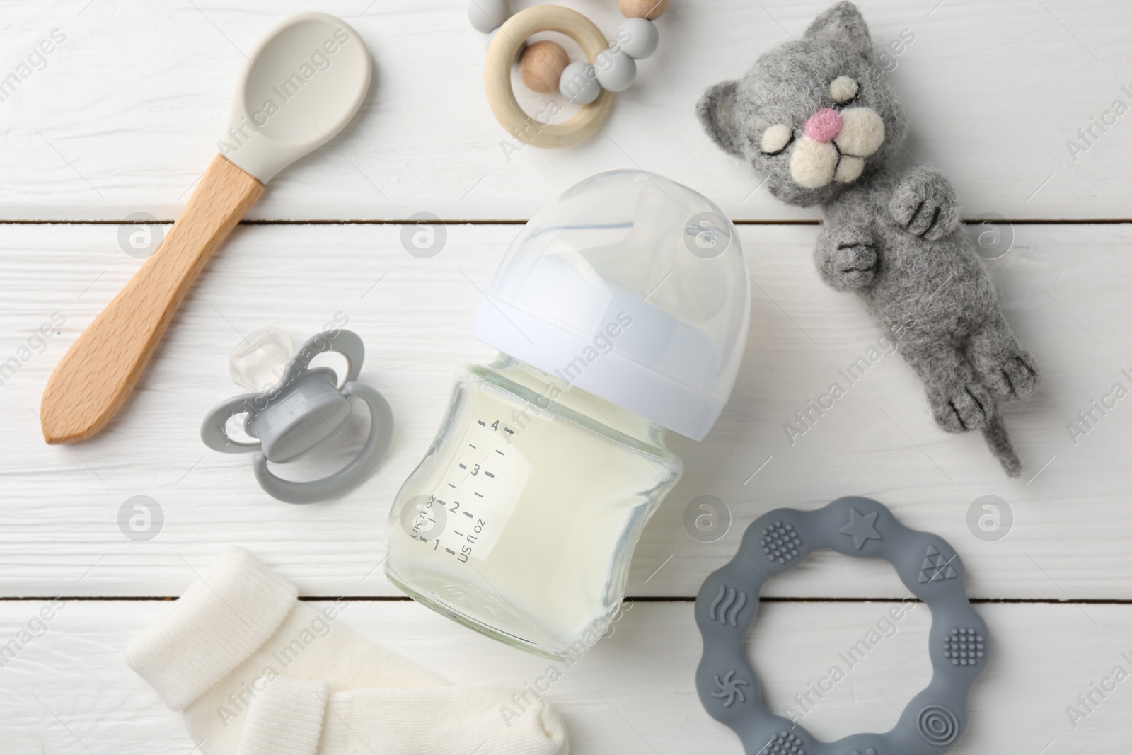 Photo of Feeding bottle with milk, needle felted cat, pacifier, spoon, baby socks and teethers on white wooden table, flat lay