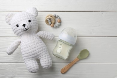 Feeding bottle with milk, toy bear, teether and spoon on white wooden table, flat lay. Space for text