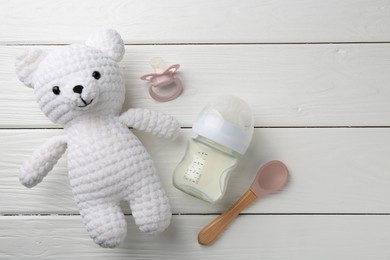 Feeding bottle with milk, toy bear, pacifier and spoon on white wooden table, flat lay. Space for text