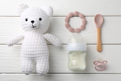 Feeding bottle with milk, toy bear, pacifier, spoon and teethers on white wooden table, flat lay