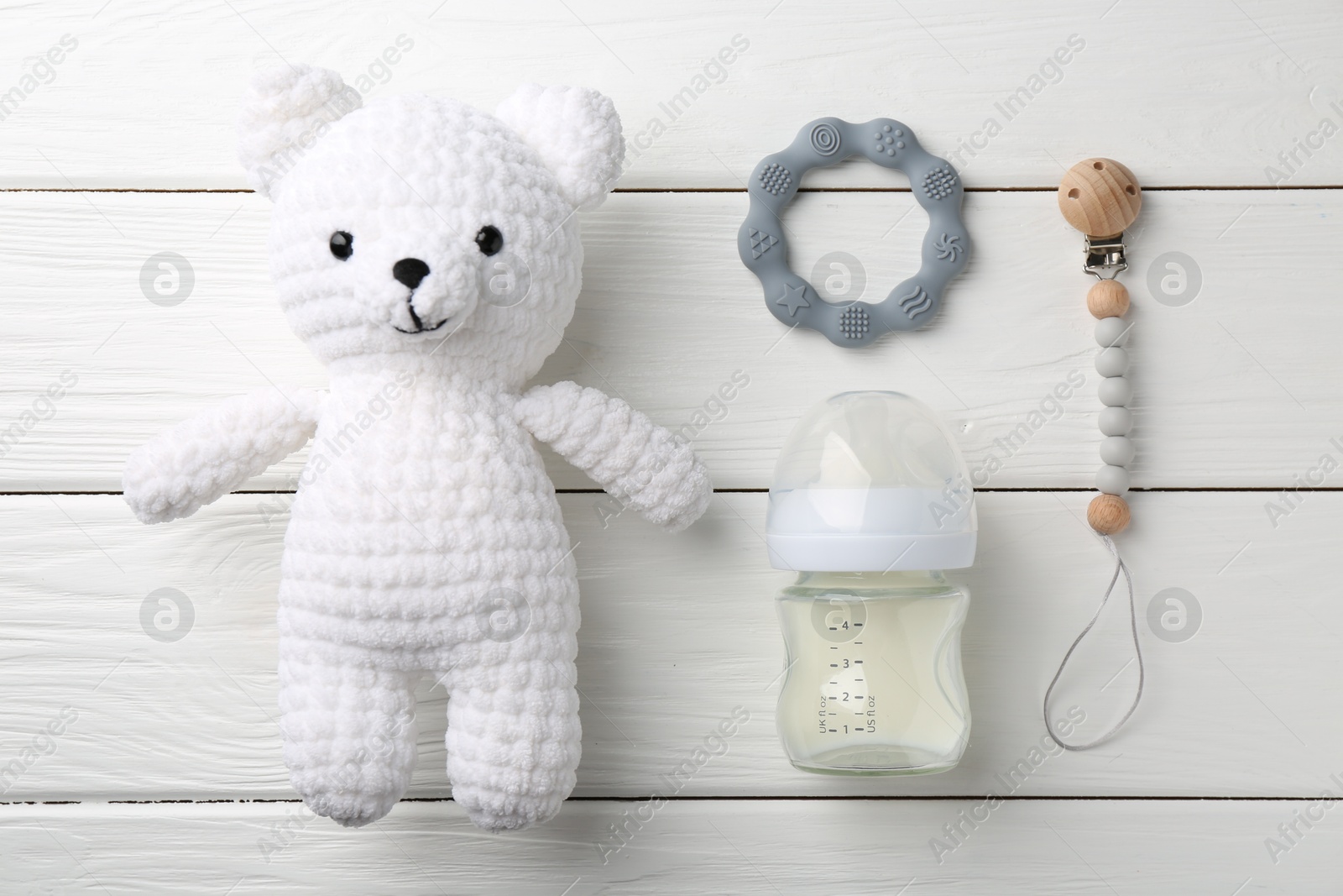 Photo of Feeding bottle with milk, toy bear and teethers on white wooden table, flat lay