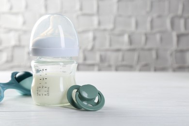 Feeding bottle with milk, pacifier and baby rattle on white wooden table, space for text