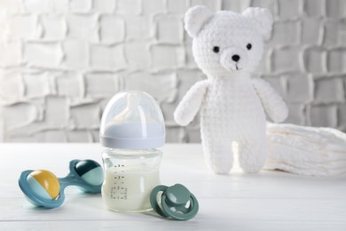 Feeding bottle with milk, baby rattle, pacifier, toy bear and diapers on white wooden table