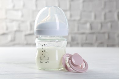 Feeding bottle with milk and pacifier on white wooden table