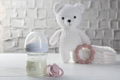 Feeding bottle with milk, pacifier, toy bear, diapers and teether on white wooden table