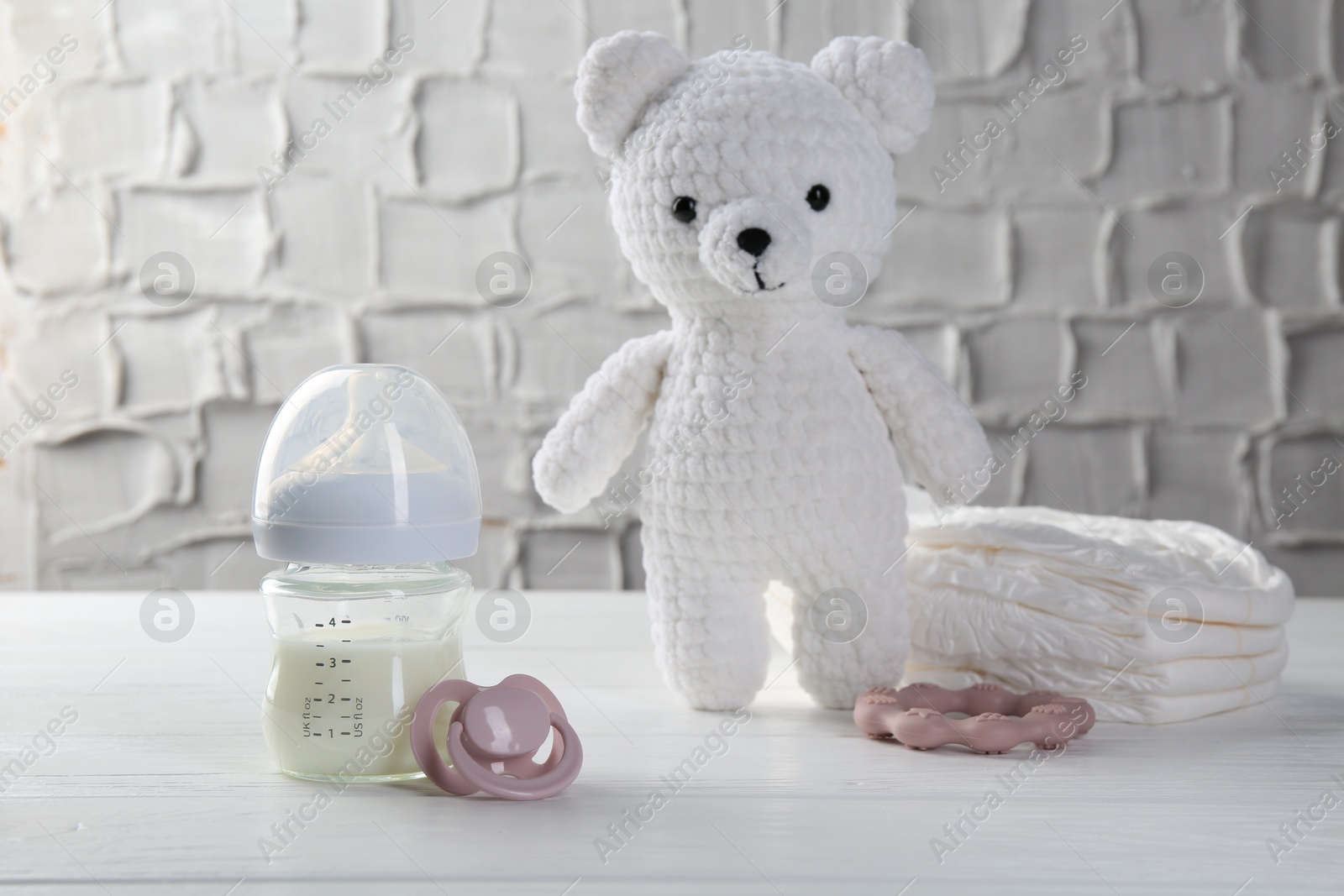 Photo of Feeding bottle with milk, pacifier, toy bear, diapers and teether on white wooden table