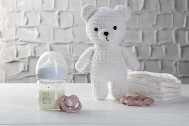 Feeding bottle with milk, pacifier, toy bear, diapers and teether on white wooden table