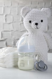 Feeding bottle with milk, toy bear, diapers and pacifier on white wooden table