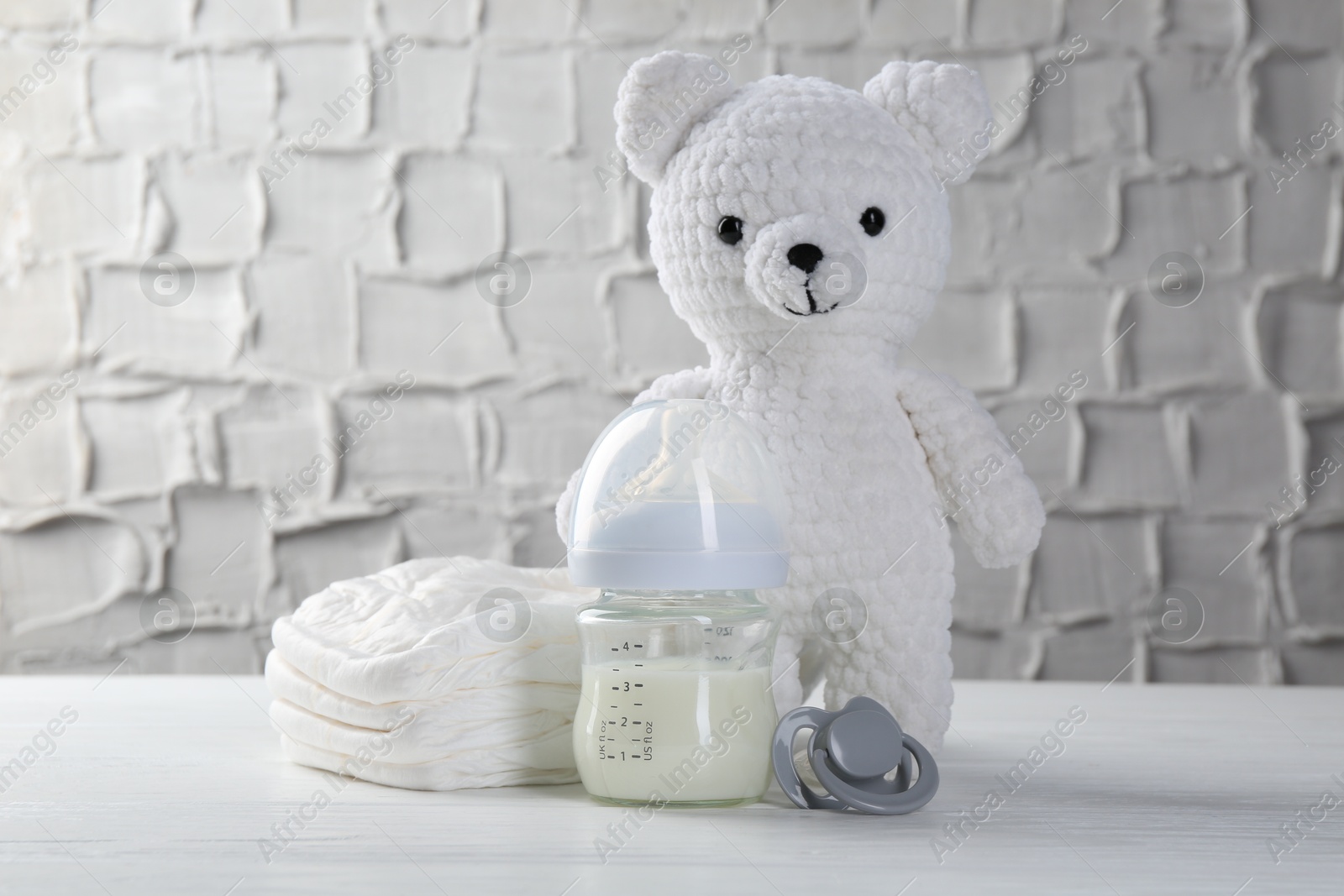 Photo of Feeding bottle with milk, toy bear, diapers and pacifier on white wooden table