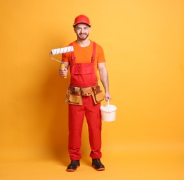 Professional painter with roller and bucket of paint on orange background