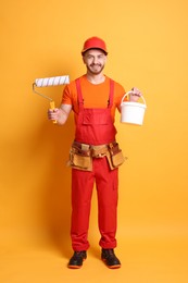 Professional painter with roller and bucket of paint on orange background