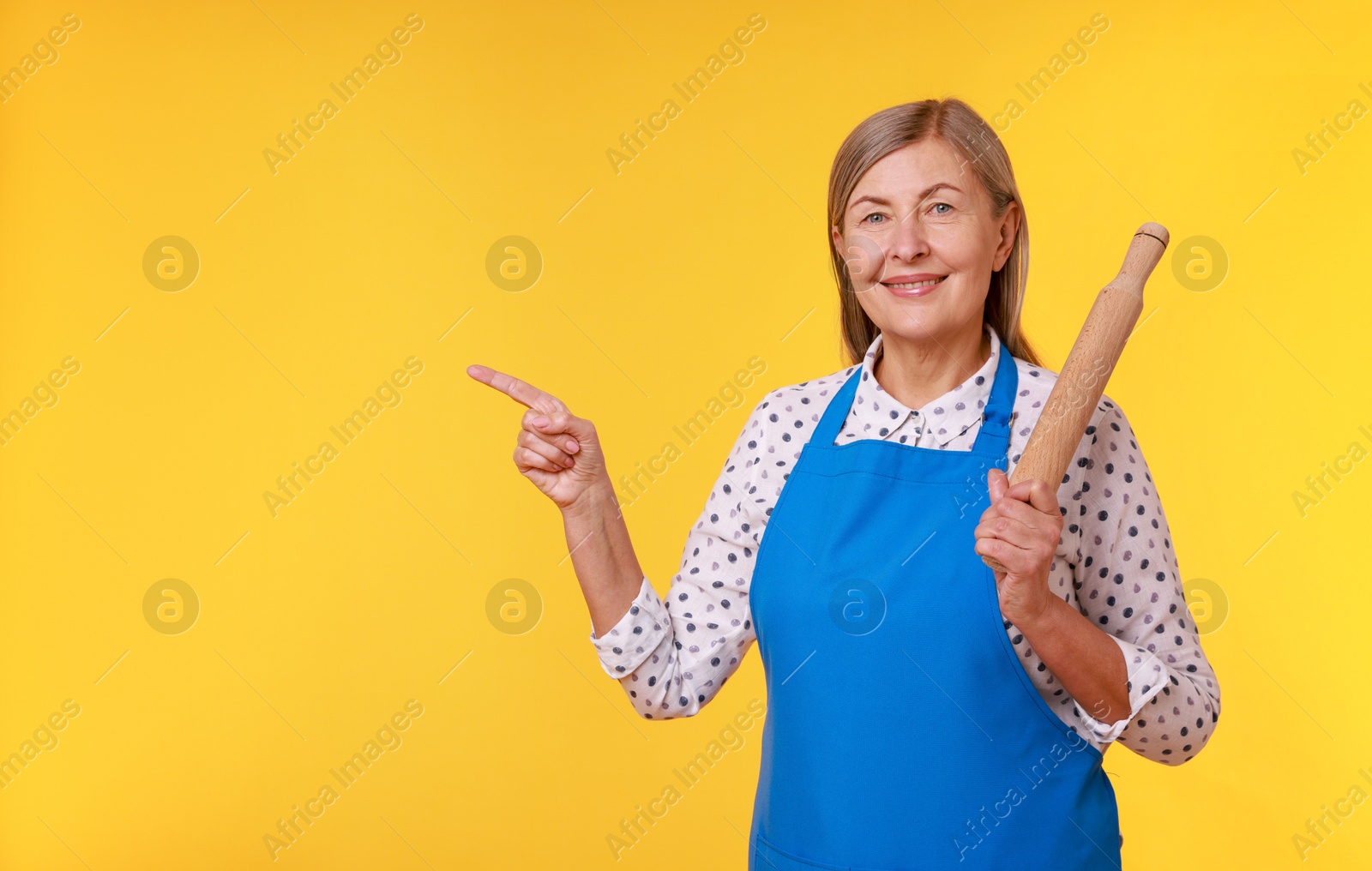 Photo of Happy woman with rolling pin pointing at something on yellow background. Space for text