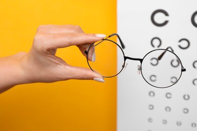 Photo of Woman holding glasses against vision test chart on orange background, closeup