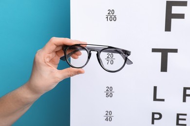 Photo of Woman holding glasses against vision test chart on light blue background, closeup