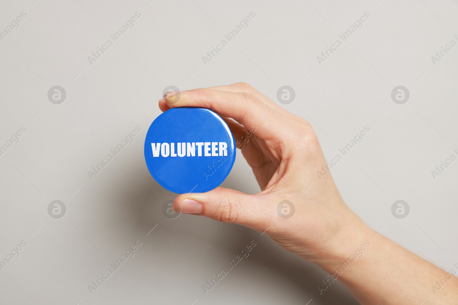 Photo of Woman holding button badge with word Volunteer on grey background, closeup