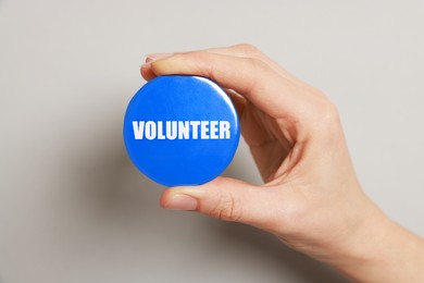 Woman holding button badge with word Volunteer on grey background, closeup