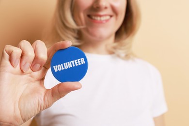 Photo of Woman holding button badge with word Volunteer on beige background, closeup