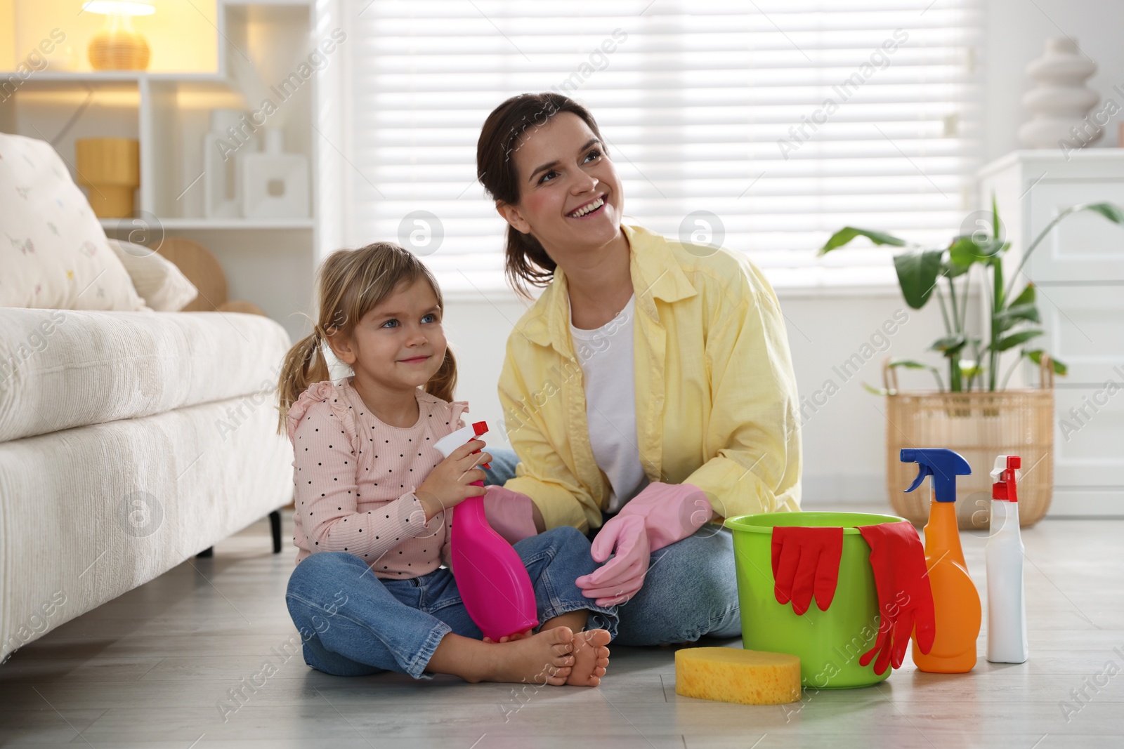 Photo of Little helper. Mother and her cute daughter with spray bottle of cleaning product on floor at home