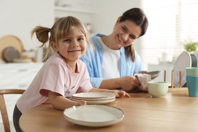 Photo of Little girl helping her mom wiping plates at table in kitchen