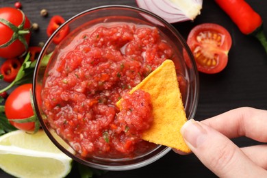 Photo of Woman dipping nacho chip into spicy salsa sauce at table, top view