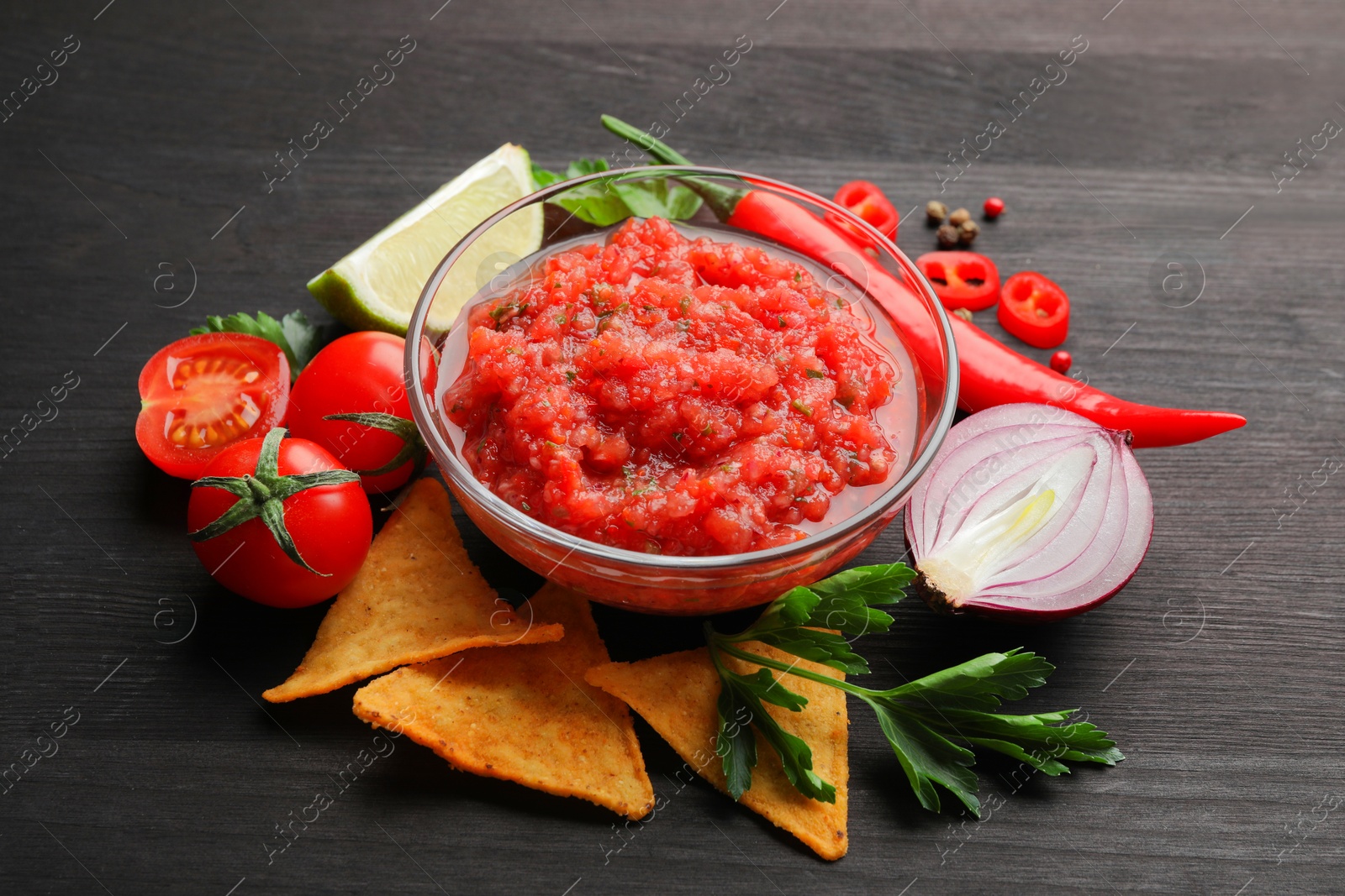 Photo of Spicy salsa sauce in bowl, nachos and ingredients on wooden table, closeup