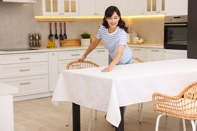 Photo of Young woman putting white tablecloth on table in kitchen