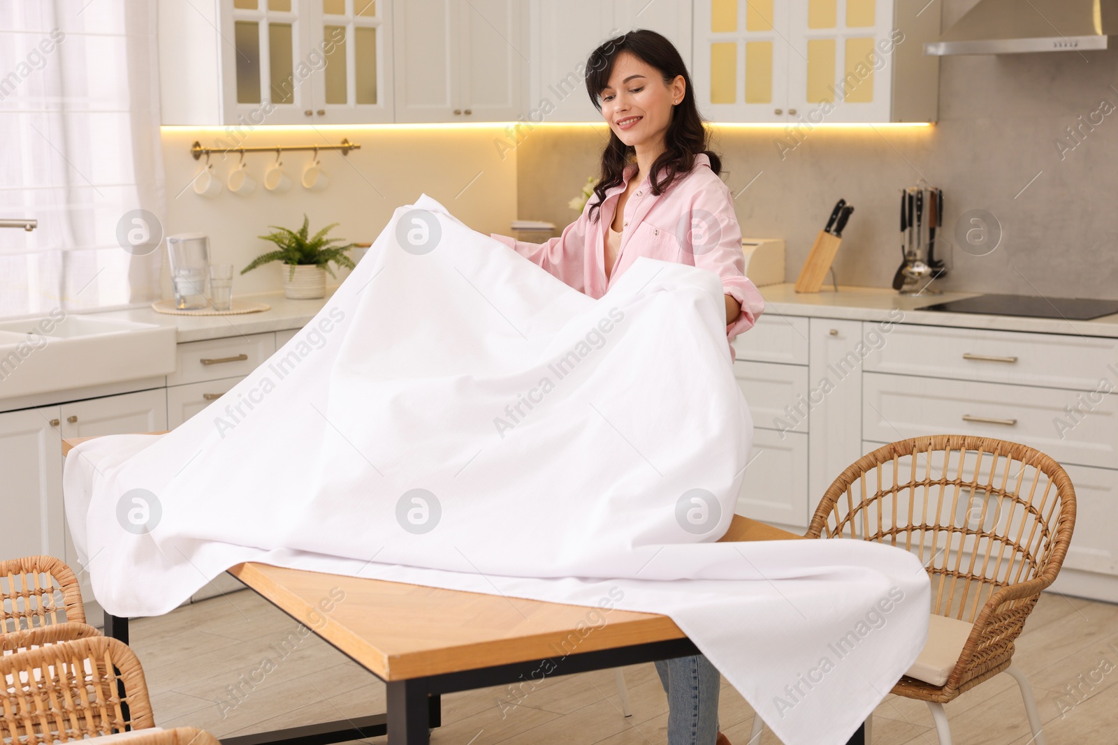 Photo of Young woman putting white tablecloth on table in kitchen