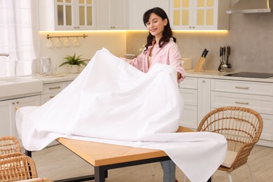 Young woman putting white tablecloth on table in kitchen