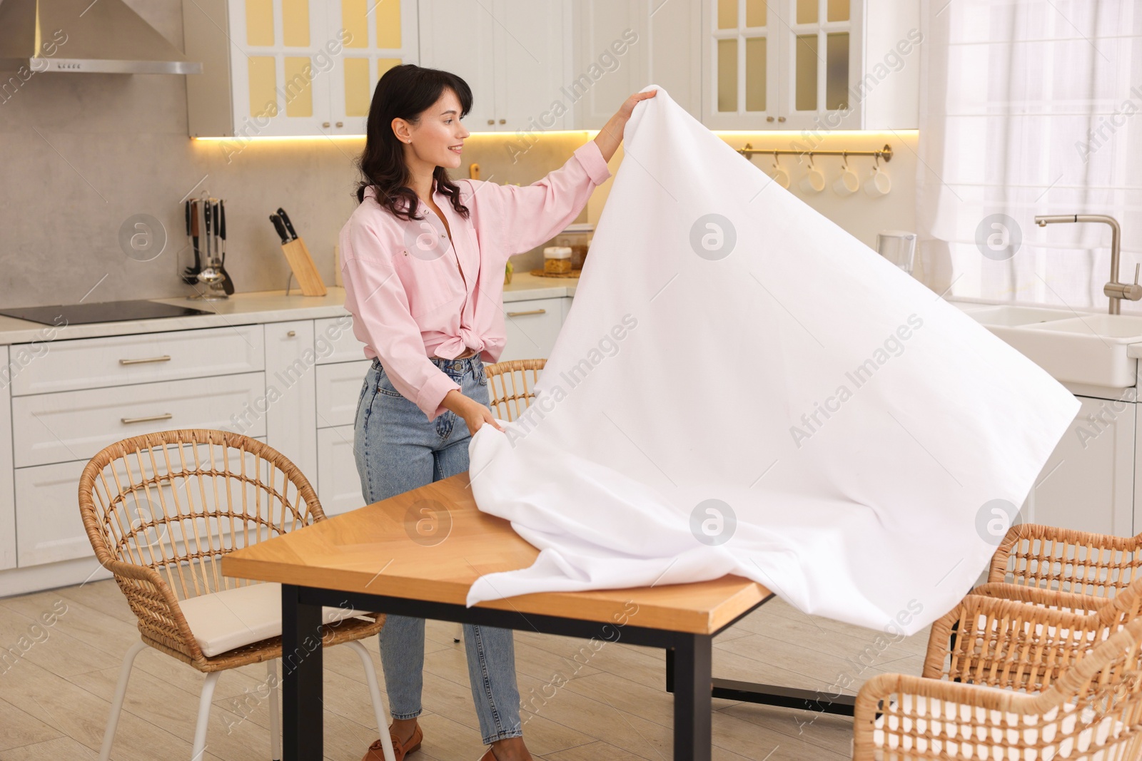 Photo of Young woman putting white tablecloth on table in kitchen