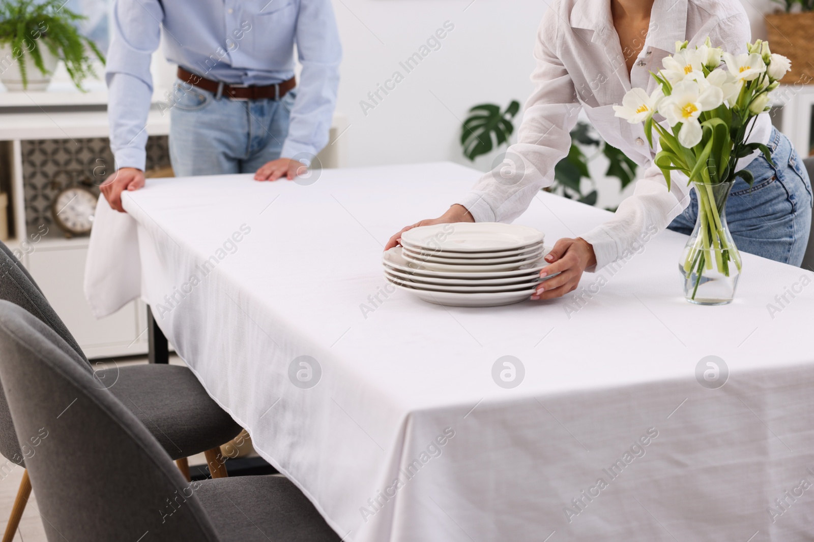 Photo of Couple putting plates on table with white tablecloth at home, closeup