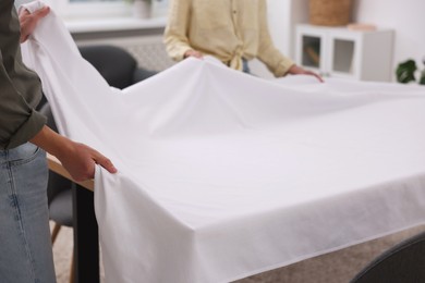 Photo of Couple putting white tablecloth on table at home, closeup