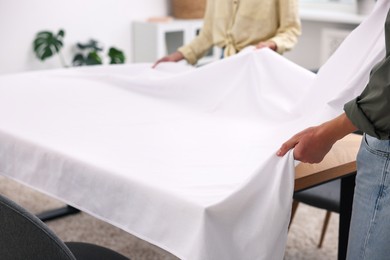 Photo of Couple putting white tablecloth on table at home, closeup