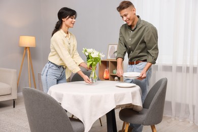 Couple putting plates and vase with flowers on table with white tablecloth at home